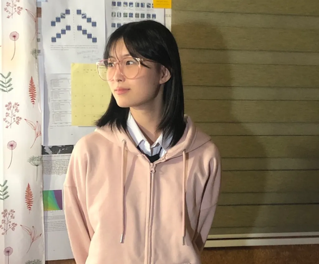 허쉬 역할의 배우 유가은이 촬영 장소인 컨테이너 내부에 분홍 후드와 안경을 쓰고 서 있는 모습.